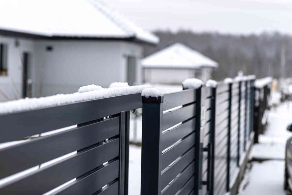 Snow covers a modern gray fence, with houses and a snowy landscape in the background.