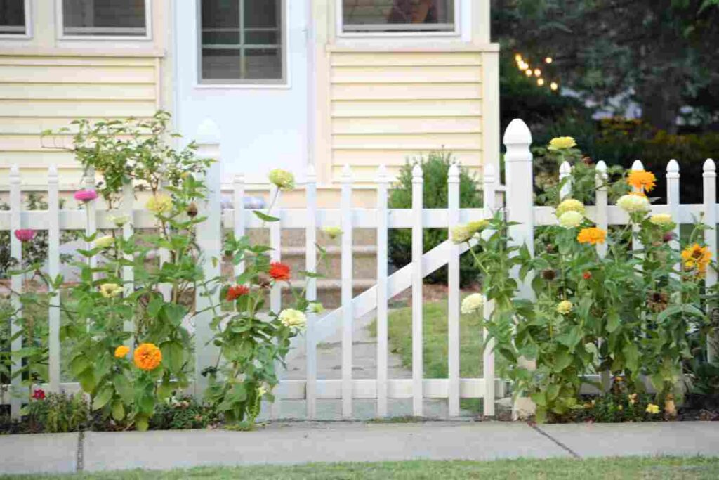 Colorful flowers grow beside a white picket fence, leading to a beige house with a door.